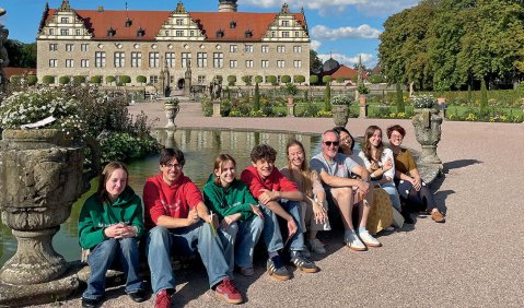 Eine Gruppe junger Menschen vor einem Schloss unter blauem Himmel.