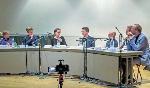 Halbrund zusammengestellte Tische mit den Menschen dahinter sitzend.