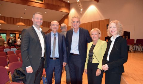 5 Menschen (im Schnitt im höheren mittleren Alter) stehen in schicker Kleidung in einer Art Stadthalle.