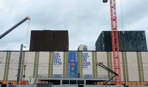 Eine Gebäude hinter sehr hohem Bauzaun und zwischen Kränen.