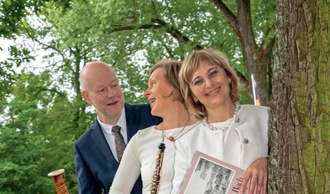 Drei Menschen im mittleren Aler stehen hinter einen Baum im Freien in Konzertkleidung. Oboistin und Fagottist haben ihre Instrumente in der Hand.