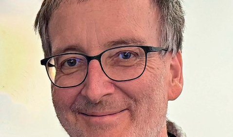 Ein Mann höheren mittleren Alters mit kurzen grauen Haaren, Brille und grauem Hemd und Pullover. Er lächelt zurückhaltend und herzlich.