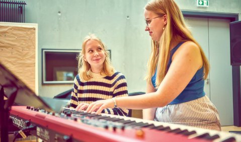 Zwei junge Frauen (eine davon eher eine Jugendliche) an einem Stage-Piano: Die eine scheint der anderen Unterricht zu geben.