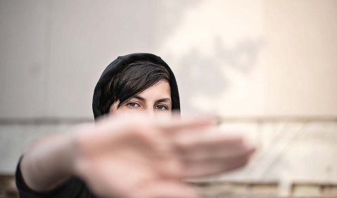 Eine Person mit Kapuze und dunklen Haaren die quer über die Stirn fallen hält die ausgestreckte Hand vor die Kamera, dass nur die Augen und Stirn des Gesichts noch zu sehen sind.
