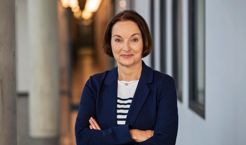 Eine Frau in mittlerem Alter mit halblangen glatten dunklen Haaren und dunkelblauem Blazer in einem Foyer.