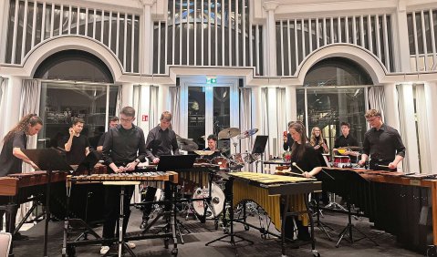 Ein großes Schalgwerkensemble spielt vor einer Wand aus vielen weißen Fensterbögen mit weißen Streben darüber.