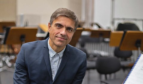 Ein Mann mittleren Alters sitzt in blauem Anzug ohne Krawatte in einem leeren Probenraum auf einem Stuhl und lächelt in die Kamera.