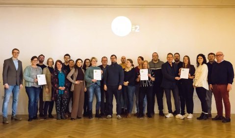 Rund zwei Dutzend Menschen überwiegend mittleren Alters stehen in einer Reihe auf einem Pakettboden vor einer Wand. 4 Personen halten exemplarisch ihre Schulungsbeschneinigung in den Händen.