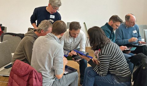 Mehrere Personen (teils sitzend, teils stehend) stecken buchstäblich die Köpfe zusammen während sie auf eihrem Schoß an digitalen Endgeräten arbeiten.