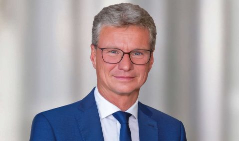 Ein Mann in höherem mittleren Alter mit kurzen grauen Haaren, angerundeter rechteckiger Brille in einem blauen Anzug mit blauer Krawatte.