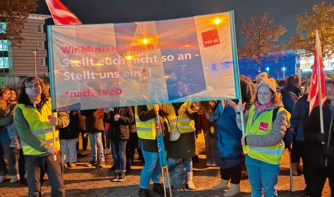 Eine Gruppe von Menschen in Warnwesten und mit roten ver.di-Flaggen und großen Transparenten/Plakaten zu dunkler Stunde in orangenem Straßenlaternenlicht.