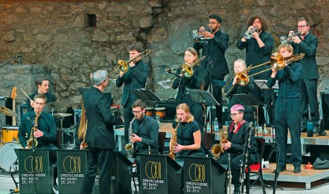 Eine Bühnen aus mehreren gestaffelten Podesten vor einer mittelalterlichen Steinmauer. Auf der Bühne eine junge Bigband in schwarz.
