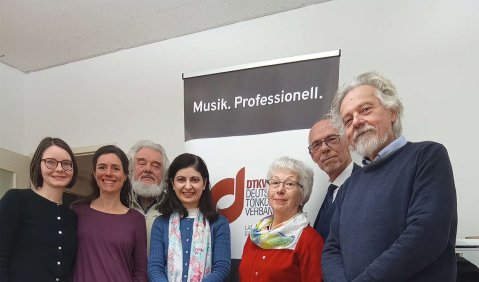 Mehrere Männer und Frauen in unterschiedlichen Altersgruppen stehen beisammen in einem gefliesten Raum.