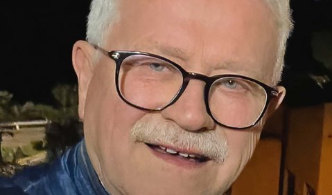 Ein Mann im höheren mittleren Alter mit weißen Haaren und Bart und schwarzer größerer abgerundeter Brille. Er trägt eine blaue Daunenjacke und lächelt in die Kamera.