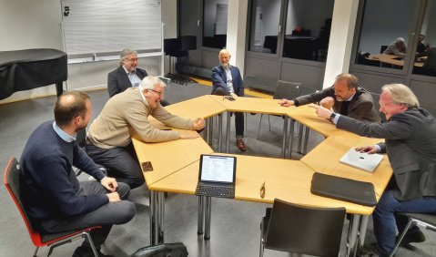 Mehrere zu einem Kreis zusammen geeschobene Tische, daran sitzen mehrere Männer überwiegend mittleren bis höheren mittleren Alters zusammen. Der Tisch steht in einem musikalischen Seminarraum.