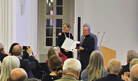 Ein voller Saal. Vorne stehen drei Menschen mit Dokumenten.