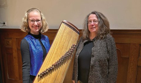 Eine junge und eine ältere Frau stehen in einem mit Parkett ausgelegten und teils holzvertäfelten Raum. die Jüngere hält ein an ein Hackbrett erinnerndes Instrument in die Kamera.