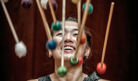 Eine Frau mit schwarzem Kleid steht hinter vielen fliegenden und bunt gewickelten Mallet-Schlägeln 