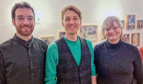 Zwei jüngere Männer und eine etwas ältere Frau vor einer Wand mit Fotos.