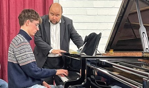 Ein Klavierstudent wird von einem neben ihm stehenden Mann mit halbglatze instruiert.