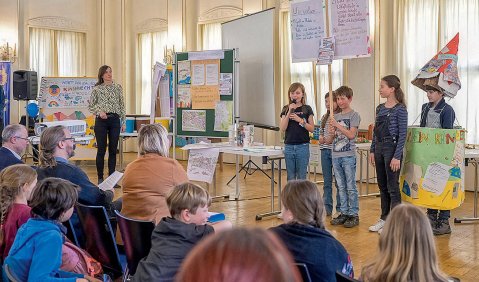 In einem alten Saal führt eine Gruppe von Kindern mit Kostümen uind Instrumenten etwas vor. Links steht eine Frau. Vor den Kindern sitzen einige Erwachsene und Kinder im Publikum.