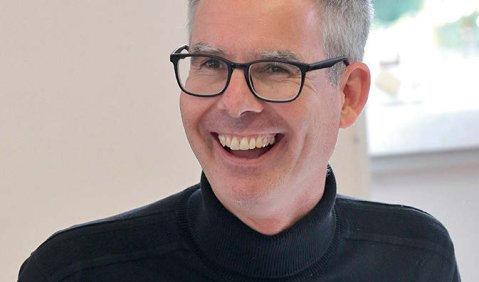 Ein Mann im schwarzen Oberteil mit einer schwarzen Brille und kurzen grauen Haaren der lachend fotografiert worden ist.