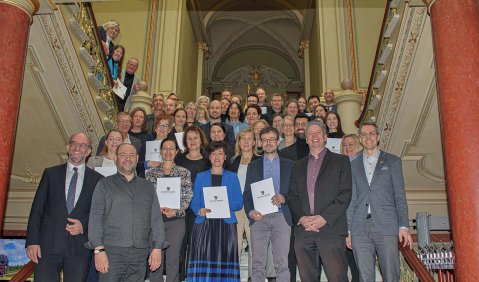 Eine größere Gruppe von Menschen (schätzungsweise 50) im mittleren Alter steht auf einer breiten Treppe eines Altbaus zu einem Gruppenfoto zusammen.