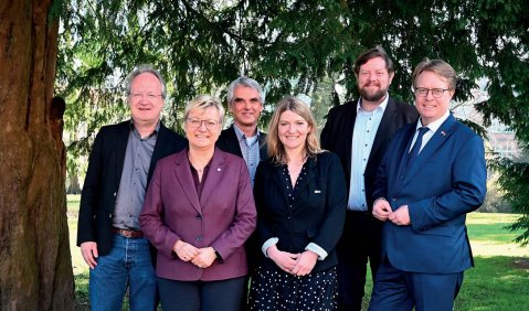 Die sechs Personen stehen (in gehobener Kleidung) unter einem Baum für ein Foto bereit.
