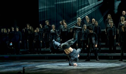 Wirkt wie ein Foto einer besonderen Operninszenierung: In dunklem bläulichen Licht breakt ein Breakdancer vor der Kulisse eines großen klassischen Chores und einer aus gräulich-bläulichen Rückwand mit einem Muster, das entfernt an Orgelpfeifen erinnert.