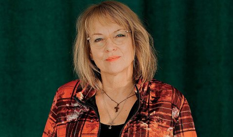 Eine rotblonde Frau in einem rot-schwarz gemusterten Blazer vor einem schwarzen Hintergrund. Sie hat schulterlanges Haar und trägt orange-roten Lippenstift.