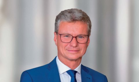 Ein Mann mit grauen Haaren und Brille in blauem Anzug vor einer weißgestreiften Wand.