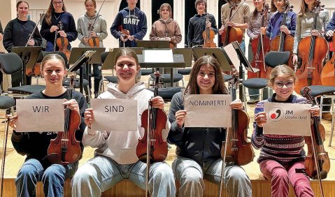 Ein Streichorchester. Kinder in der ersten Reihe halten ausgedruckte Worte hoch: "Wir sind nominiert!" und das Logo der Jeunesses Musicales.
