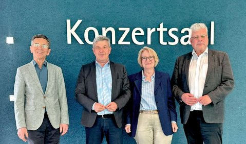 Gruppenfoto mit den vier Personen. Sie tragen alle Blazer beziehungsweise Sakko. Im Hintergrund eine blaue Wand mit der Aufschrift Konzertsaal in großen weißen Buchstaben.