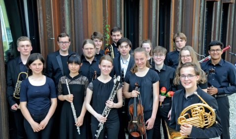Gruppenfoto mit mehreren Jugendlichen mit Instrumenten vor einer gestreiften Wand.