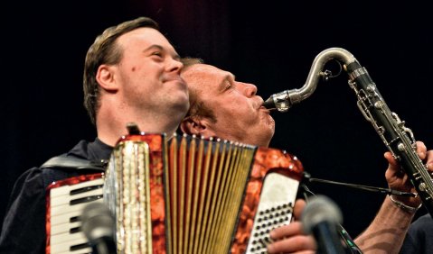 Zwei Musiker spielen aneinander gelehnt vor einem schwarzen Hintergrund. Einer hat ein Akkorderon, er andere eine Bassklarinette oder ein schwarzes Saxofon. Vor beiden stehen zwei Mikrofone. Der Akkordeonist hat Gesichtszüge, die an das Down-Syndrom erinnern.