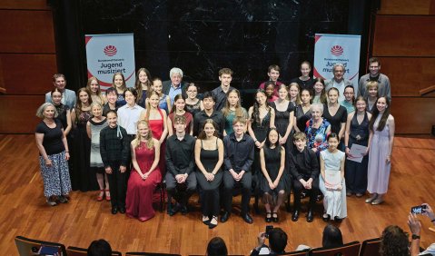 Ein Saal mit Parkett. In der Mitte sitzen und stehen viele Kinder und Jugendliche in Abendkleidung für ein gemeinsames Gruppenfoto. Flankiert werden sie von Jugend musiziert-Transparenten und ein paar Erwachsenen.