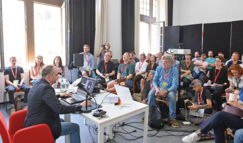 Ein mittelgroßer, aber voll beserzter Raum mit interessierten Zuhörer:innen und einem Redner mit reichlich Elektronik auf seinem Pult.