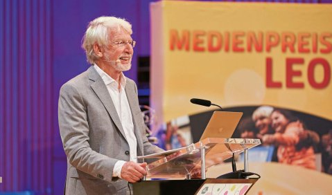 Ein Mann mit weißen Haaren und kurzem weißen Vollbart steht in einem grauen Anzug vor einem Rednerpult.