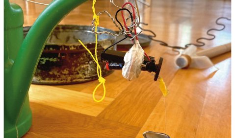 Nahaufnahme verschiedener verbundener Gegenstände: Eine Gießkanne, einige Schnüre, Steine, Kabel, eine Pfanne und das Ende eines Besenstiels. Alles steht auf dem Boden oder hängt an der Gießkanne. Im Hintergrund zwei Menschen.