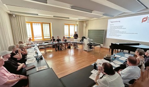 Etwa 20 Personen sitzen an einem Rechteck aus Tischen mit einem Flügel und einer Leinwand an einer Seite für eine Konferenz beisammen.
