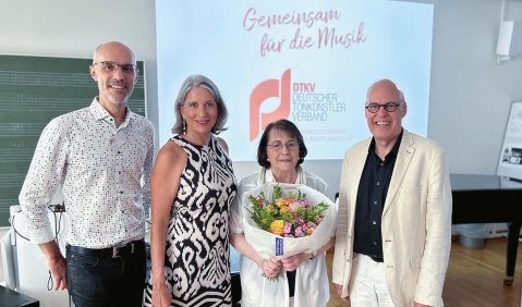 Vier Erwachsene im höheren mittleren Alter in feierlicher heller Kleidung. Im Hintergrund ein projiziertes DTKV-Logo