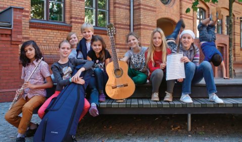 Mehrere Kinder sitzen mit Instrumenten auf einer Bank und einem Tisch auf dem Schulhof vor einer Schule. Die Kinder haben verschiedene Instrumente dabei.