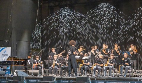 Eine Bigband vor schwarz-weißem Hintergrund beim Spielen auf einer Bühne.