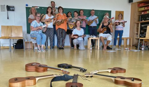 Vorne im Bild liegen Ukulelen in verschiedenen Größen in einem Kreis. Weiter hinten in dem Raum sitzt und steht eine kleine Menge älterer Menschen mit Ukulenen.