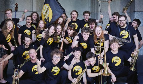 Viele Jugendliche in schwarzen T-Shirts mit gelbem Kreis jubeln mit ihren Instrumenten (Bigband-Instrumente)