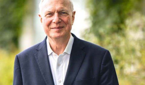 Ein älterer Mann mit sehr kurzer weißer Halbglatze steht in Anzug vor einem grünen Hintergrund und lächelt freundlich in die Kamera.