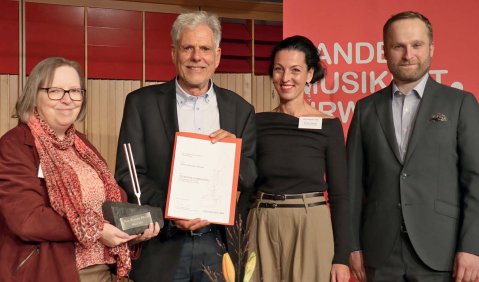 Vier Personen stehen zu einem Foto zusammen. Hans Hermann Wickel trägt einen Anzug, kurzgeschnittene weiße Haare und eine Urkunde in den Händen. Ihm wird eine Stimmgabel auf einem Steinsockel überreicht.