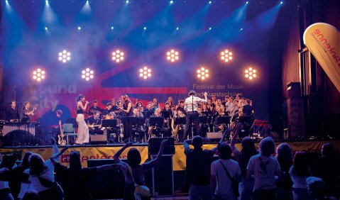Eine bunt beleuchtete Bühne mit mehreren Lichtern die die Form von quasi stilisierten Blumen haben. Davor musizieren teils stehend teils sitzend eine Bigband mit vielen Sänger:innen.