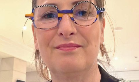 Nahes Portrait einer Frau mittleren Alters mit runder bunter Brille (obere Hälfte Blau-weiß-getigert, unten Orange Blau)