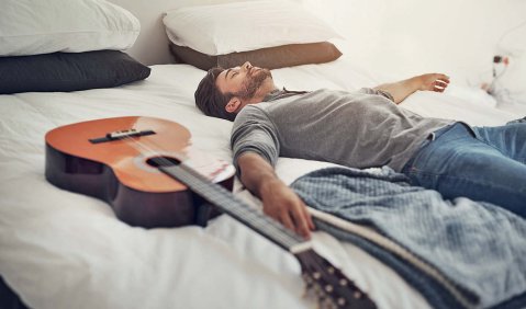Ein Gitarrist liegt neben seiner Gitarre in Straßenklamotten auf einem gemachten Bett.